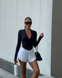 Woman in black long-sleeve top and white shorts standing against a light-colored wall.