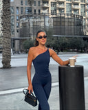 Woman in a navy blue one-shoulder jumpsuit holding a coffee cup and a black handbag, standing in an urban setting.